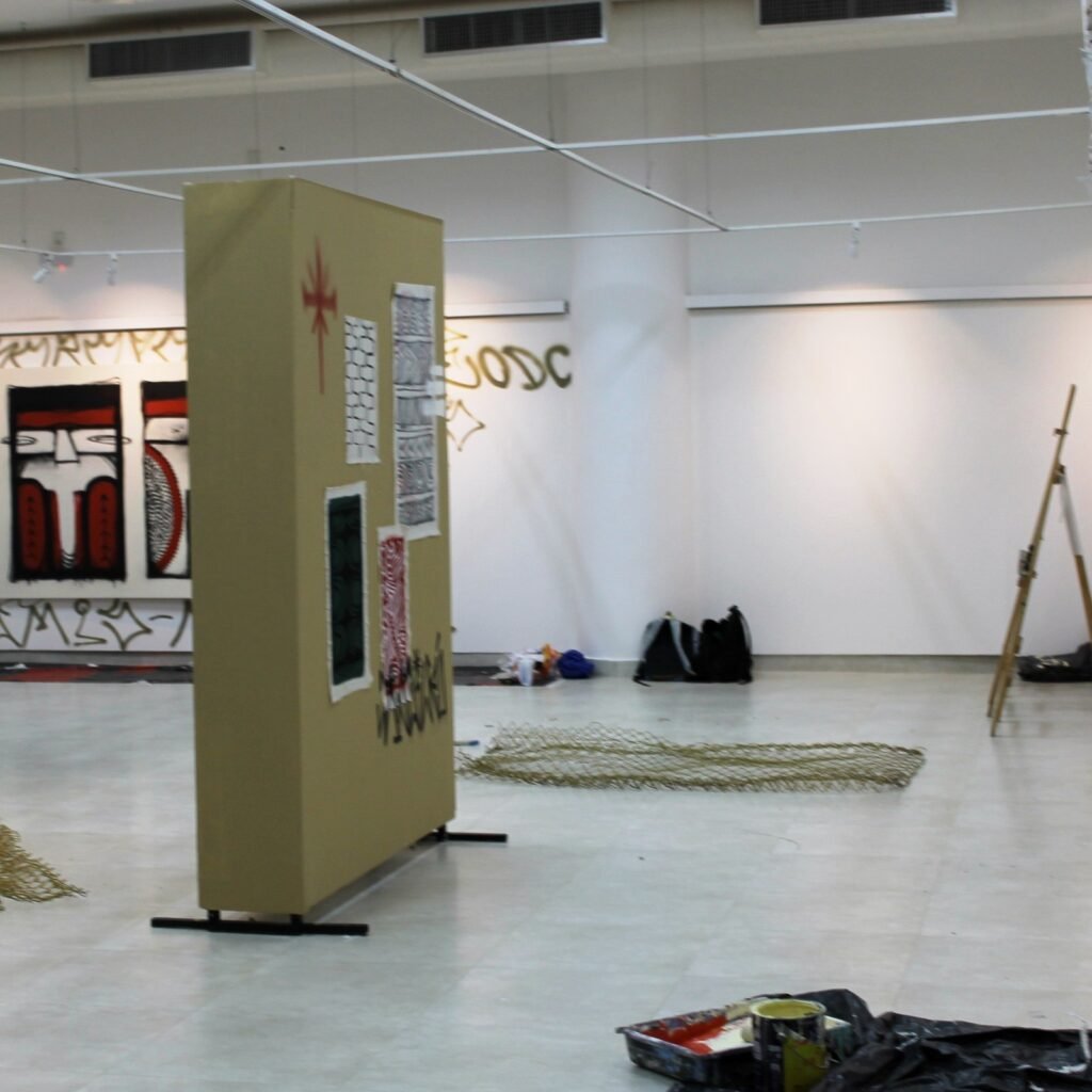 Sala de exposiciones durante el montaje: panel a medio instalar, caballete, lona y botes de pintura en el suelo