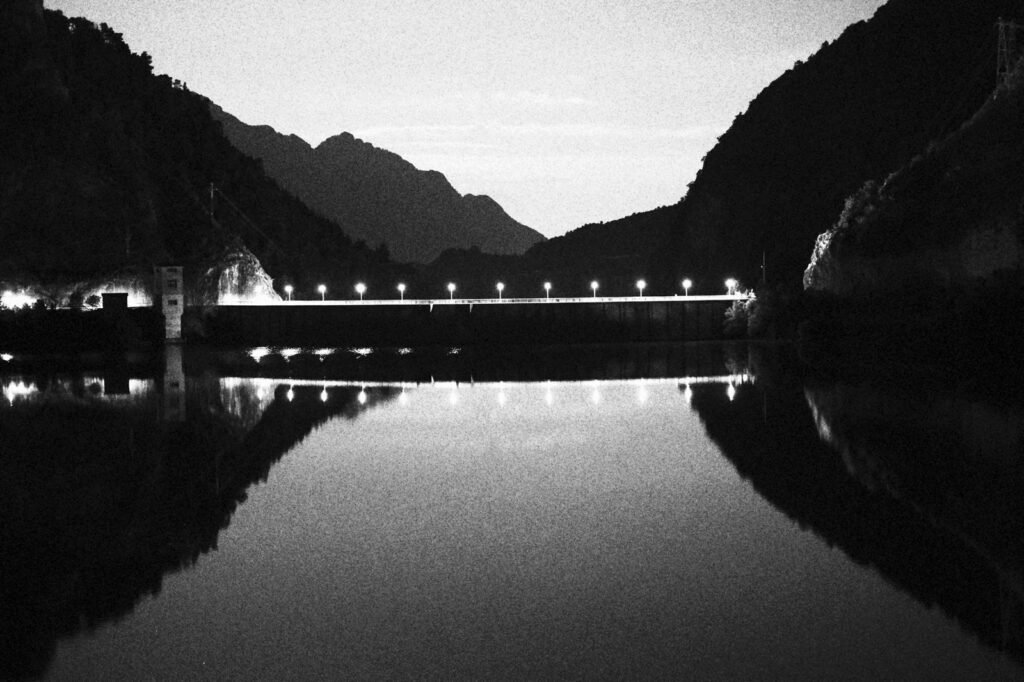 Embalse de noche con farolas reflejadas en el agua bajo las montañas, en blanco y negro